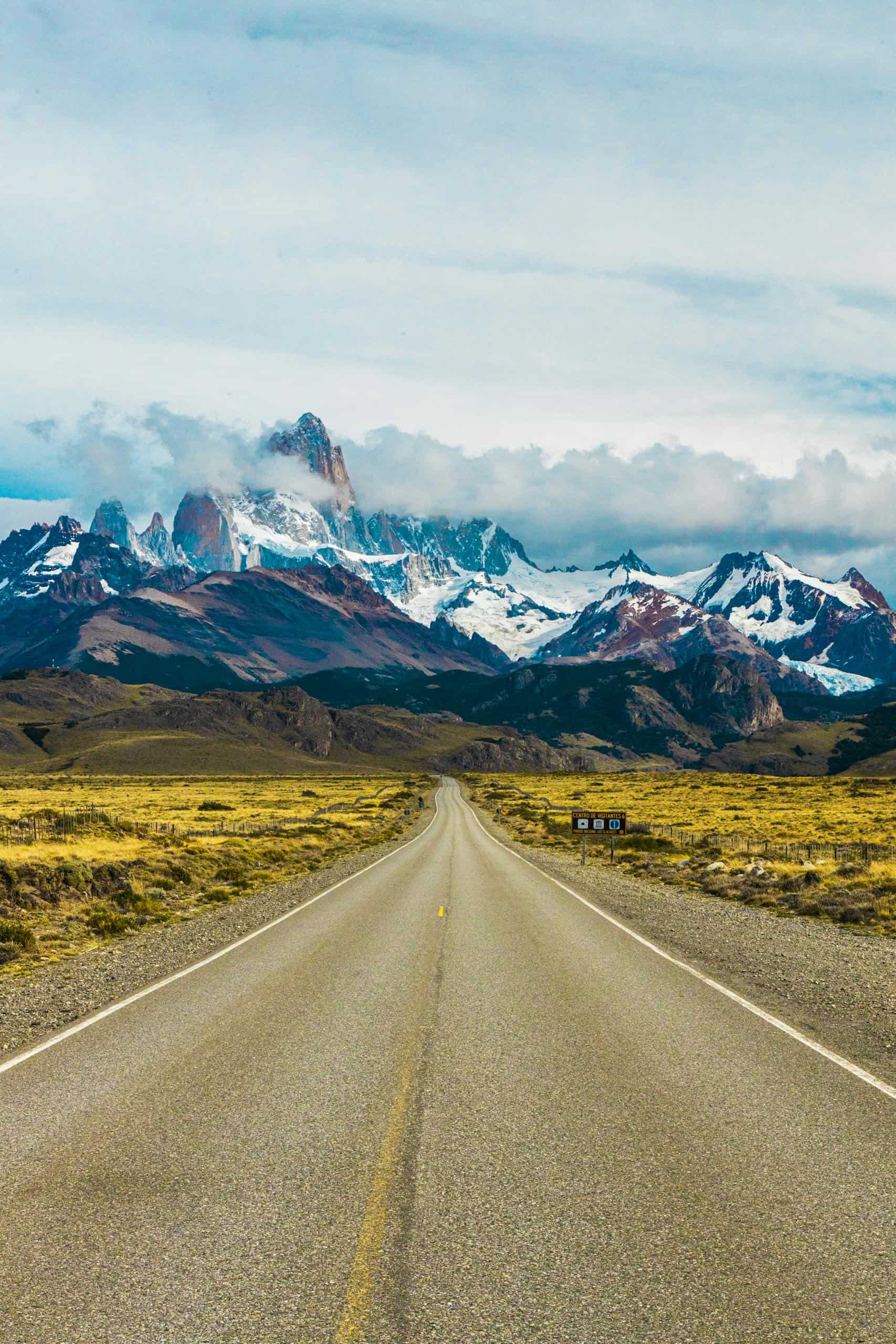 Long stretch of road leading to spectacular snow-capped mountains under a clear blue sky.