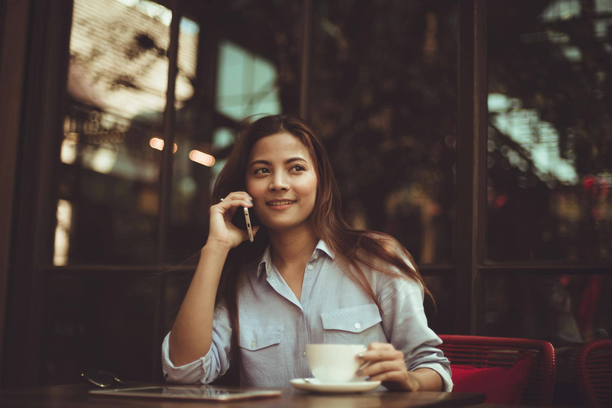 Young woman in casual shirt talking on phone and holding coffee at an outdoor café table.
